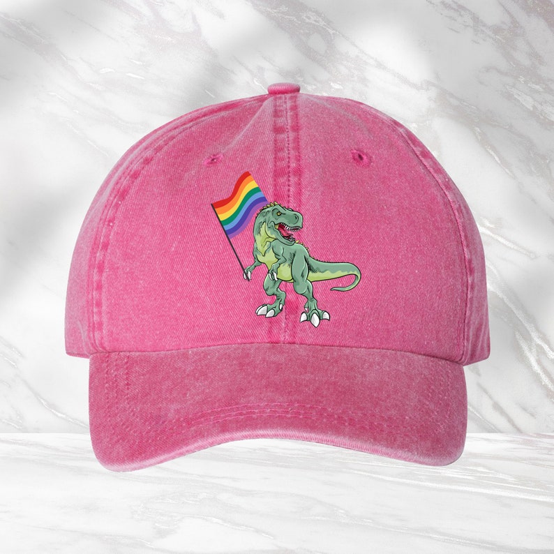 Puede incluir: Gorra de b&eacute;isbol rosa con un dinosaurio verde sosteniendo una bandera arco&iacute;ris.