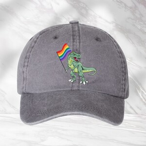 Puede incluir: Una gorra de b&eacute;isbol de denim gris con un dinosaurio verde sosteniendo una bandera arco&iacute;ris del orgullo.