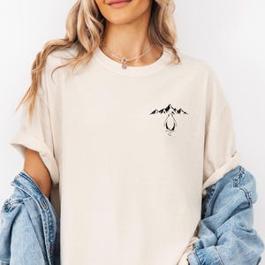 A woman wearing a white t-shirt with a graphic design of mountains and a penguin on the front. She is also wearing a denim jacket over the t-shirt.