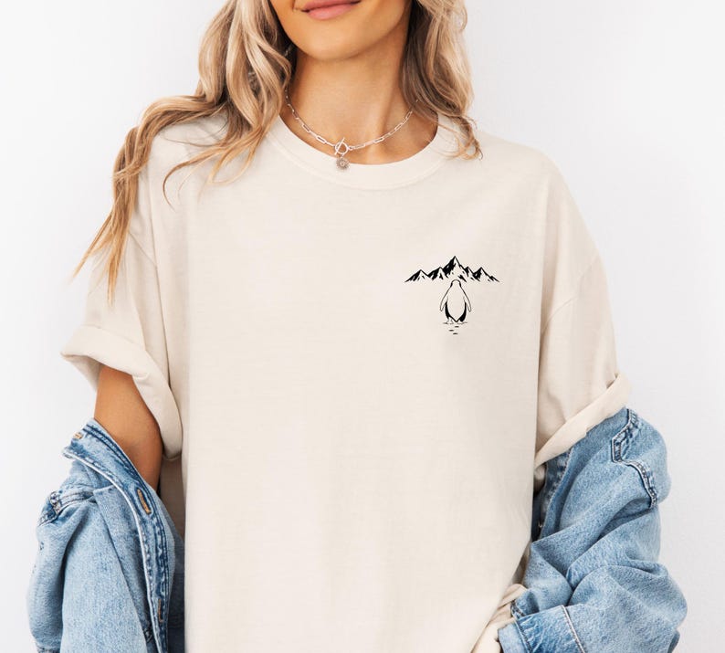 A woman wearing a white t-shirt with a graphic design of mountains and a penguin on the front. She is also wearing a denim jacket over the t-shirt.