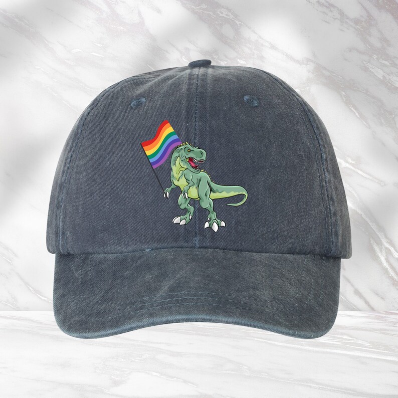 Puede incluir: Una gorra de b&eacute;isbol de denim azul oscuro con un dinosaurio verde sosteniendo una bandera arco&iacute;ris del orgullo.