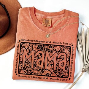 A pink t-shirt with a floral design and the word "Mama" printed on it, along with the text "Motherhood is Kingdom Work - Proverbs 31:25-27" and "Motherhood is Kingdom Work - Proverbs 31:25-27" printed around the edges. The t-shirt is placed on a white surface, with a brown hat and a plant visible in the background.