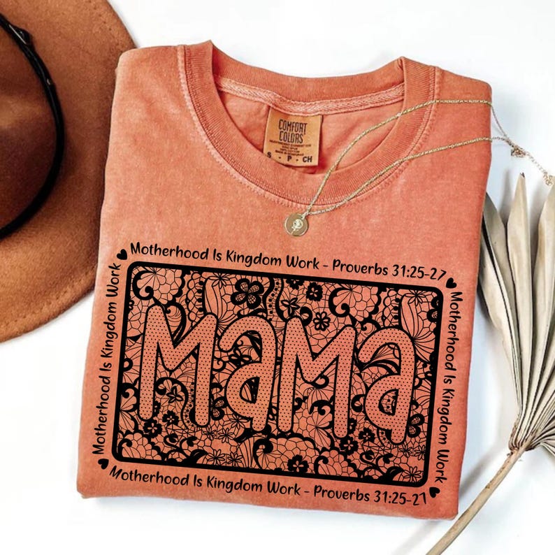 A pink t-shirt with a floral design and the word "Mama" printed on it, along with the text "Motherhood is Kingdom Work - Proverbs 31:25-27" and "Motherhood is Kingdom Work - Proverbs 31:25-27" printed around the edges. The t-shirt is placed on a white surface, with a brown hat and a plant visible in the background.