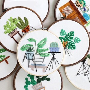 May include: A set of six embroidery hoops with various plant designs, including cacti, ferns, and monstera plants. The hoops are made of wood and have a white background. The embroidery is done in green, brown, and white thread.