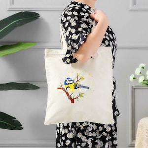 May include: A white canvas tote bag with a bluebird embroidered on it. The bird is perched on a branch with yellow flowers. The bag has long handles.