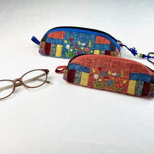 May include: Two fabric eyeglass cases with embroidered floral designs. The top case is blue with a red, yellow, and blue patchwork design. The bottom case is red with a blue, yellow, and red patchwork design. Both cases have a blue zipper and a blue and green tassel.