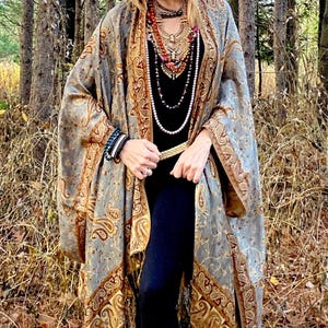 Könnte beinhalten: Eine Frau trägt einen langen, grauen und goldenen Paisley-Schal mit Fransen. Sie trägt ein schwarzes Oberteil und eine schwarze Hose. Sie ist mit einem goldenen Gürtel, mehreren Halsketten und Armbändern ausgestattet.