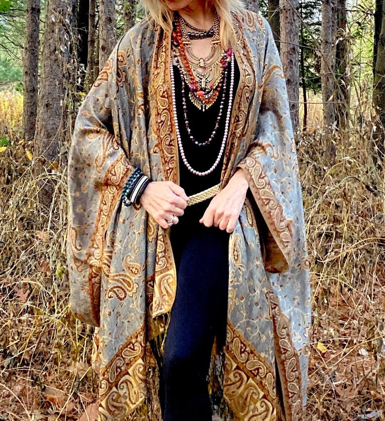 Op de afbeelding: Een vrouw draagt een lange, grijze en gouden paisley-gekleurde sjaal met franjes. Ze draagt een zwart topje en een zwarte broek. Ze is geaccessoiriseerd met een gouden riem, meerdere kettingen en armbanden.