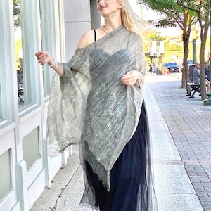 Puede incluir: Una mujer lleva un poncho gris y blanco transparente sobre una falda de tul negra. El poncho tiene un corte holgado y fluido y está hecho de una tela ligera. La falda es larga y fluida, y la mujer la lleva con un par de sandalias plateadas.