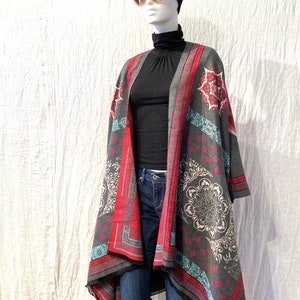 May include: A gray, red, and blue patterned poncho with a fringed edge. The poncho is worn over a black mock neck shirt and blue jeans.