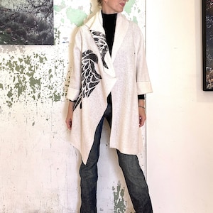 May include: A white long-sleeved cardigan with a black bird design on the front. The cardigan is open and the person wearing it is standing in front of a white wall.