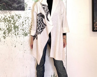 Op de afbeelding: Een witte cardigan met lange mouwen met een zwart vogelontwerp op de voorkant. De cardigan is open en de persoon die hem draagt staat voor een witte muur.