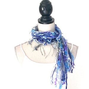 May include: A blue, white, and purple scarf with frayed edges is displayed on a white mannequin. The scarf is made of yarn and has a loose, flowing texture.