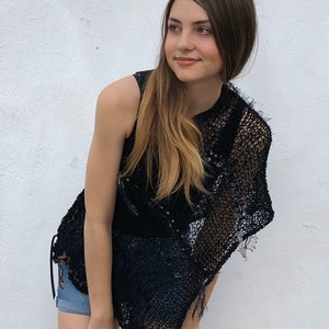 May include: A woman wearing a black crochet top with a fringe detail. The top is sleeveless and has a loose fit. The woman has long brown hair and is looking to the side.