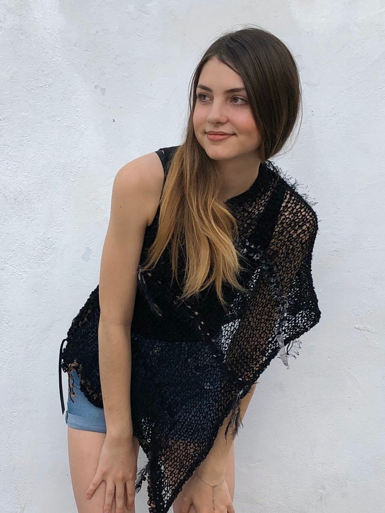 May include: A woman wearing a black crochet top with a fringe detail. The top is sleeveless and has a loose fit. The woman has long brown hair and is looking to the side.