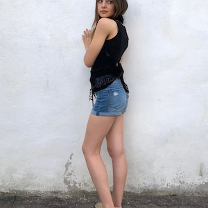 May include: A young person wearing a black sleeveless top with a crocheted design and blue denim shorts. They are standing in front of a white wall and wearing beige sneakers.