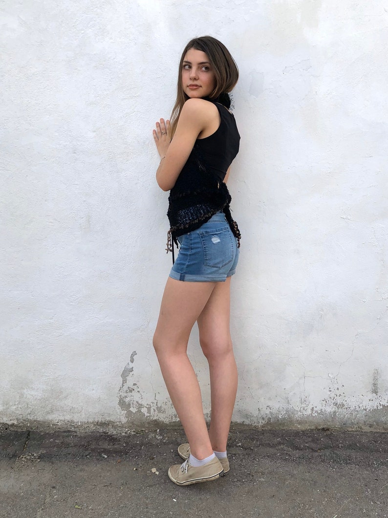 May include: A young person wearing a black sleeveless top with a crocheted design and blue denim shorts. They are standing in front of a white wall and wearing beige sneakers.