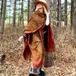 Op de afbeelding: Een vrouw draagt een lange, rood-groene paisley sjaal met een bontkraag. Ze staat in een bos en draagt een brede hoed en een zonnebril.