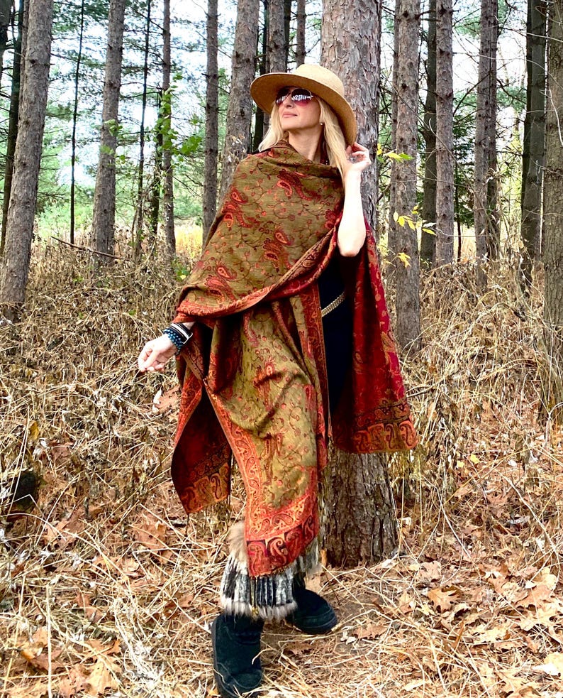 Op de afbeelding: Een vrouw draagt een lange, rood-groene paisley sjaal met een bontkraag. Ze staat in een bos en draagt een brede hoed en een zonnebril.