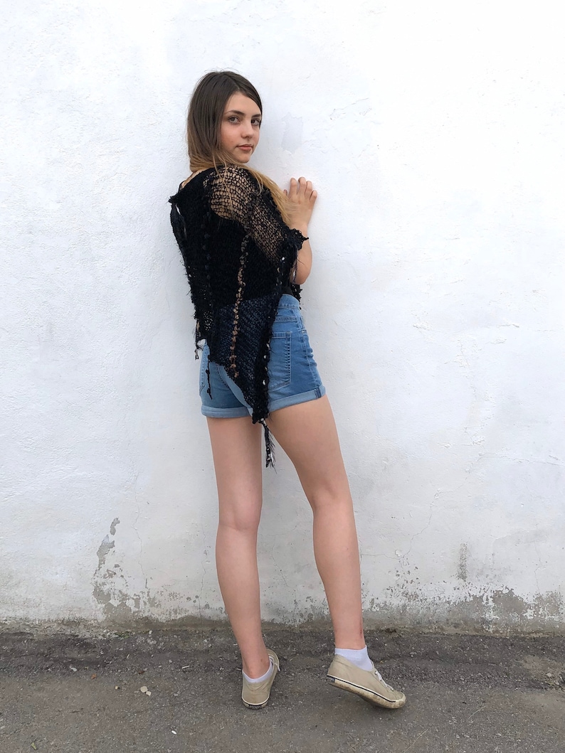 May include: A young person wearing a black crochet top and denim shorts, standing in front of a white wall. They are wearing beige sneakers.