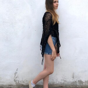 May include: A woman wearing a black crochet top with a fringe hem and denim shorts. She is standing in front of a white wall and wearing beige sneakers.