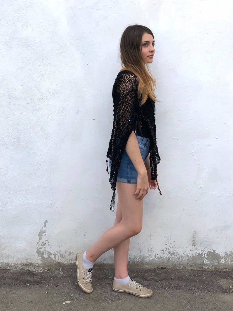 May include: A woman wearing a black crochet top with a fringe hem and denim shorts. She is standing in front of a white wall and wearing beige sneakers.