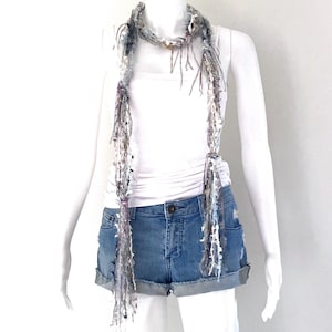 May include: A handmade, fringed scarf in shades of gray, white, and purple, draped on a mannequin. The scarf has a textured appearance with long, dangling fringe. The mannequin is wearing a white tank top and denim shorts.
