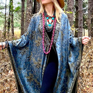 May include: A woman wearing a blue paisley patterned shawl with a long pink beaded necklace and other layered necklaces. She is standing in a woodland setting.