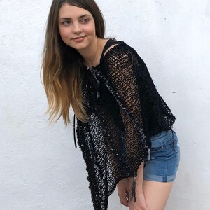 May include: A woman wearing a black crochet top with a distressed look and denim shorts. The top has a loose, flowing fit and a unique, open-weave design.
