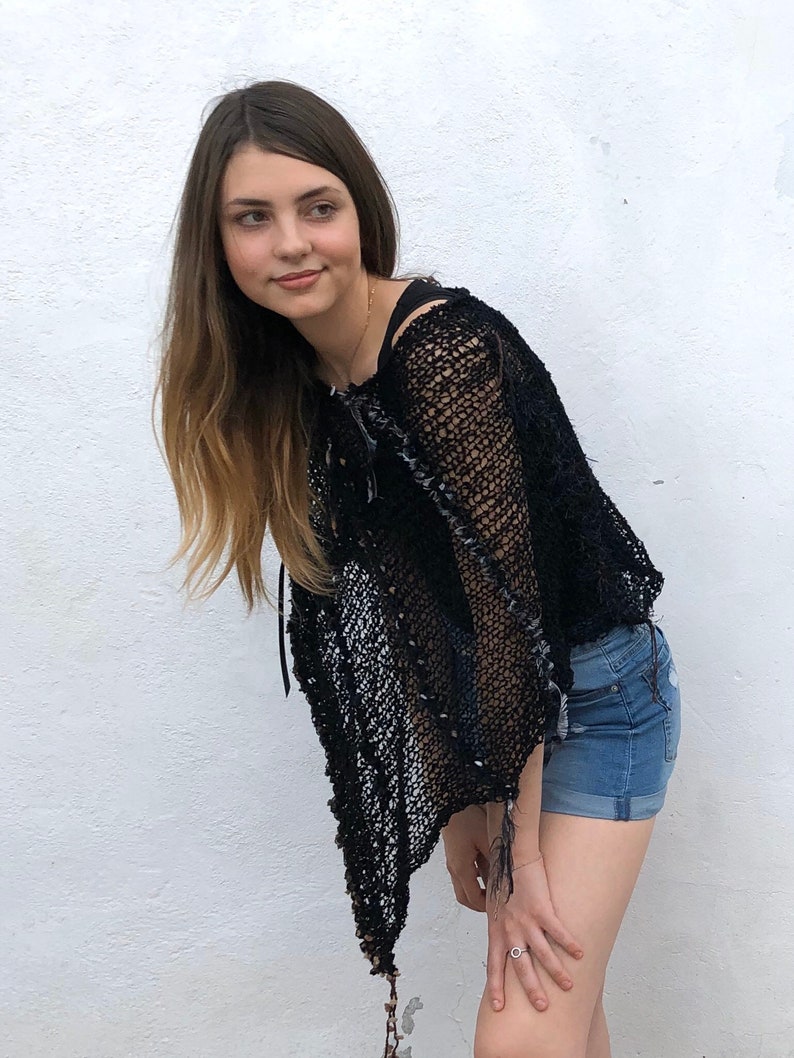 May include: A woman wearing a black crochet top with a distressed look and denim shorts. The top has a loose, flowing fit and a unique, open-weave design.