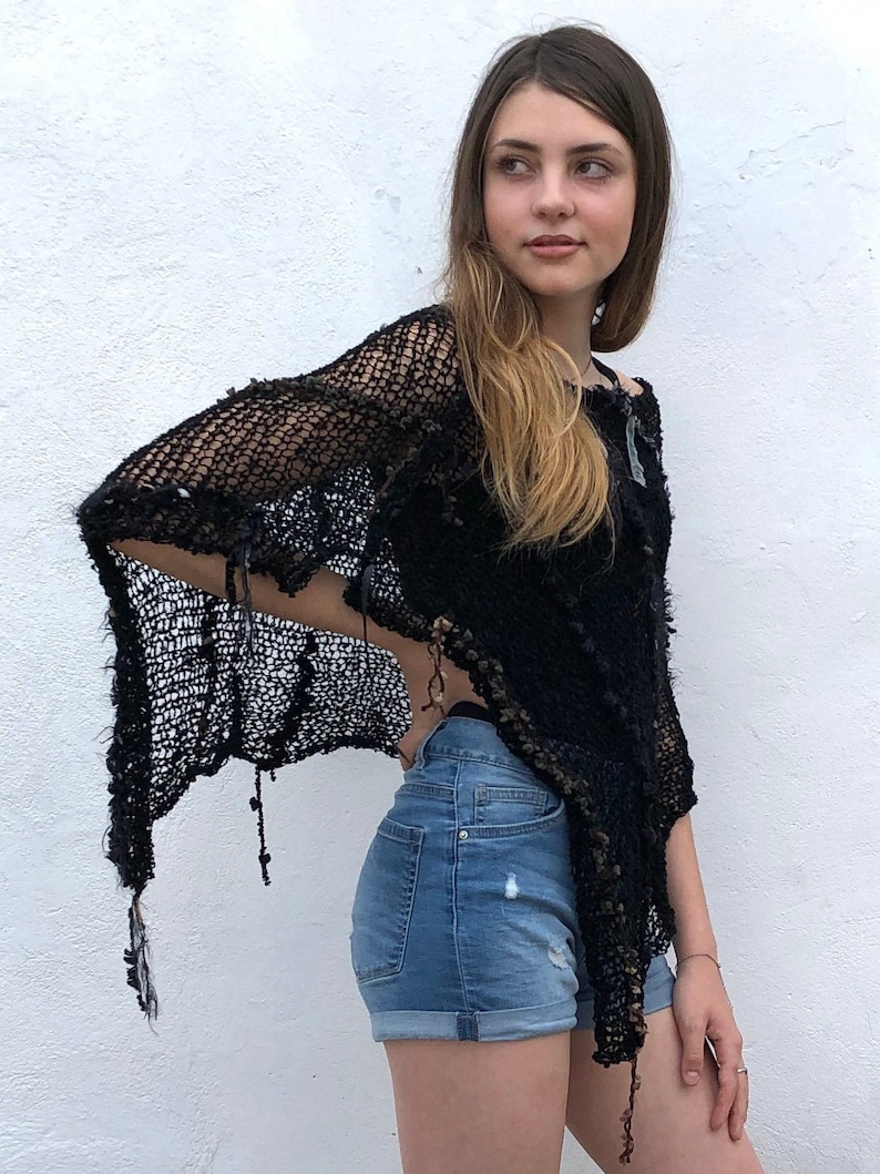 May include: A black crochet poncho with a fringe detail. The poncho is worn over a pair of blue denim shorts.