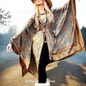 Op de afbeelding: Een vrouw draagt een lange, vloeiende, grijs-gouden paisley-sjaal met franjes. Ze draagt een brede strohoed, een zonnebril en een lange ketting met meerdere kralenstrengen. De tekst "A Sun" en "Embracing the Flow" is zichtbaar in de afbeelding.