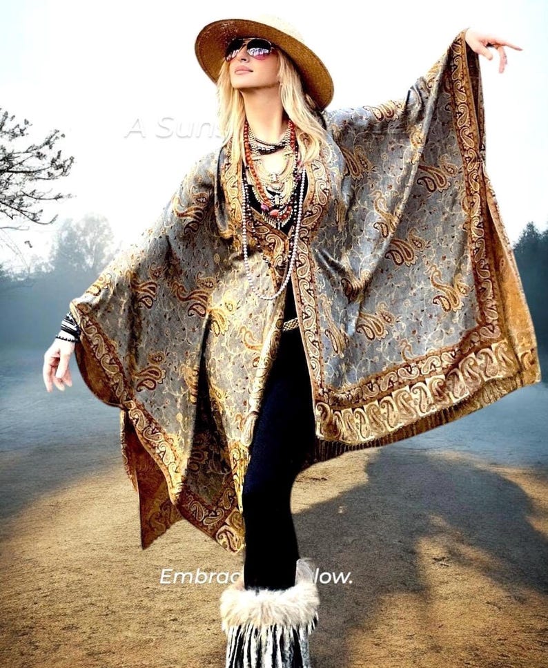 Op de afbeelding: Een vrouw draagt een lange, vloeiende, grijs-gouden paisley-sjaal met franjes. Ze draagt een brede strohoed, een zonnebril en een lange ketting met meerdere kralenstrengen. De tekst "A Sun" en "Embracing the Flow" is zichtbaar in de afbeelding.