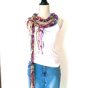 May include: A vibrant, handmade scarf featuring a mix of textures and colours, such as pink, purple, green, and blue. The scarf is displayed around a mannequin's neck, highlighting its unique design and fringed details. A fashion accessory.