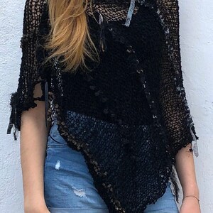 May include: Black crochet poncho with a distressed and open-weave design. The poncho is worn over a black tank top and blue denim shorts.