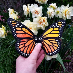 Puede incluir: Accesorio de alas de mariposa monarca con colores naranja y amarillo, ribete negro y puntos blancos. El accesorio tiene dos ojales para sujetarlo. El fondo presenta flores blancas y amarillas y follaje verde.