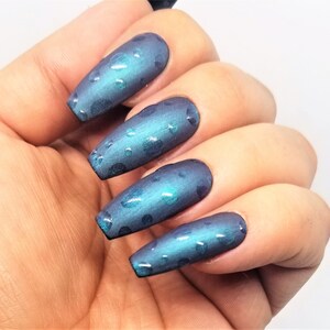 May include: Close-up of a hand with long, blue-green nails with a dark blue base and a subtle shimmer. The nails are decorated with small, light blue dots.