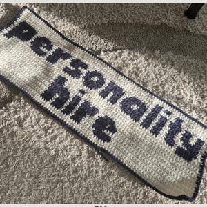 May include: A rectangular, crocheted blanket in cream with navy blue lettering that reads "personality hire". The blanket has a navy blue border and is lying on a textured, light-colored surface.