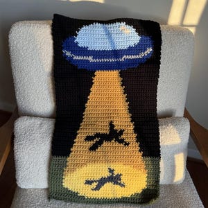 May include: A handmade crochet blanket featuring a UFO design. The blanket depicts a blue and white UFO with a yellow beam shining down on two black silhouettes. The background is black, with a green border.