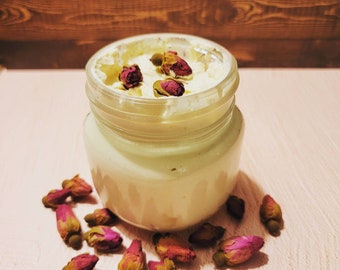May include: A glass jar filled with a creamy white substance, topped with dried rose buds. The jar is surrounded by more dried rose buds on a light pink surface.
