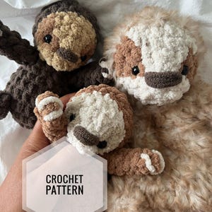 May include: Three crocheted sloth plushies in brown, tan, and white. The plushies are made with a fluffy yarn and have black eyes and brown noses. The plushies are arranged in a pile on a white surface. The text "CROCHET PATTERN" is visible in the image.