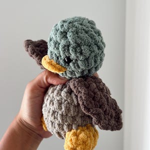 May include: A crocheted plush toy duck with a green head, brown wings, and a yellow beak. The duck is made with a soft, textured yarn.