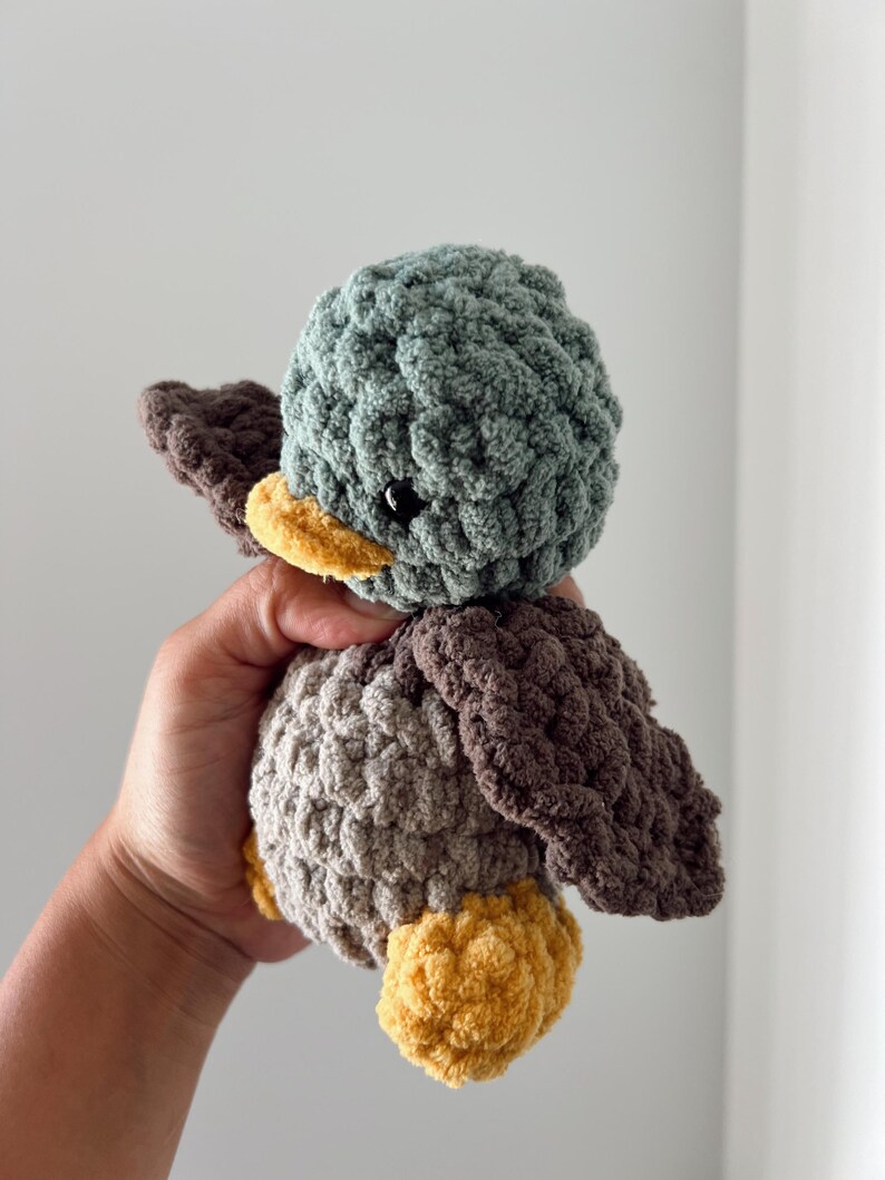 May include: A crocheted plush toy duck with a green head, brown wings, and a yellow beak. The duck is made with a soft, textured yarn.