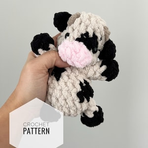 May include: Crocheted plush toy cow with black and white spots and a pink nose. The toy is made with a fluffy yarn and has a textured appearance. The text "CROCHET PATTERN" is visible in the image.
