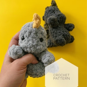May include: Two crocheted dinosaur toys, one gray with a yellow horn and one dark gray. The gray dinosaur is being held in a hand. The text "CROCHET PATTERN" is on a yellow hexagon.