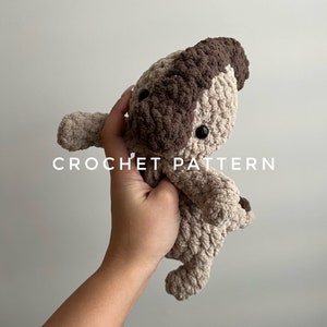 May include: A crocheted stuffed animal in the shape of a dog. The dog is brown and beige and has black eyes. The dog is being held in a hand. The text "CROCHET PATTERN" is visible below the dog.