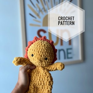 May include: A crocheted stuffed toy in the shape of a sun with a yellow body and orange hair. The toy is being held in a person's hand. The background is a light blue wall with a framed print of a sun with the word "CROCHET PATTERN" written on it.