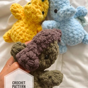 May include: Crocheted plush toys in yellow, blue, and green. The yellow toy has a green accent, the blue toy has wings, and the green and brown toy is being held. The text "CROCHET PATTERN" is in a white hexagon.