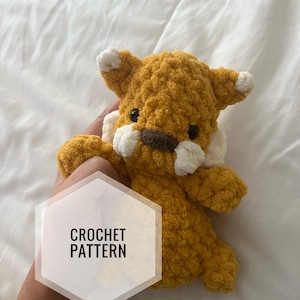 May include: Crochet pattern for a small, brown and white tiger amigurumi toy. The toy has a textured, fluffy appearance and is being held by a hand.