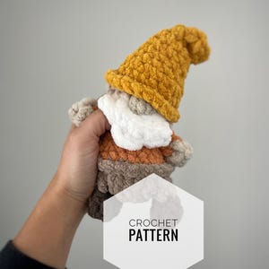 May include: Crochet pattern for a small gnome with a brown, orange, and white body and a yellow hat. The gnome is being held in a hand.